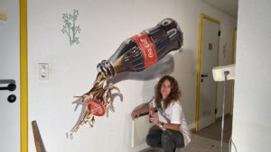 artista MaDesi davanti al Murale 3D della bottiglia di Coca Cola gignate in vetro